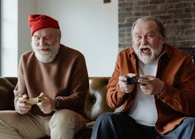 Two senior men laughing and playing video games, sharing joyful moments indoors.